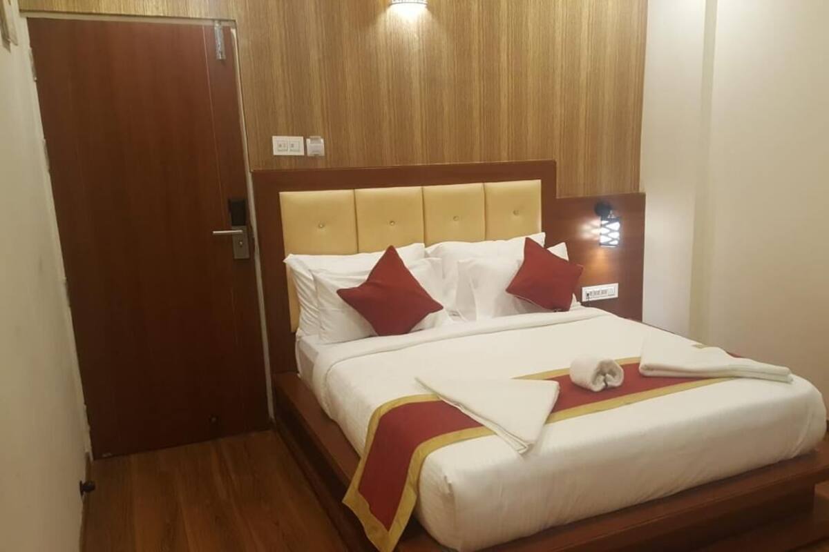 A hotel room with a wooden headboard, a bed with white bedding and red pillows, a wooden floor, and a window with curtains.