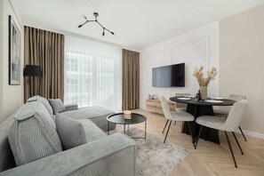 Un salon moderne avec un canapé d’angle, une table de salle à manger ronde entourée de chaises, un téléviseur à écran plat et une grande fenêtre agrémentée de rideaux.