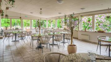 A spacious indoor dining area with multiple tables and chairs, large windows with a view of greenery, and hanging plants.