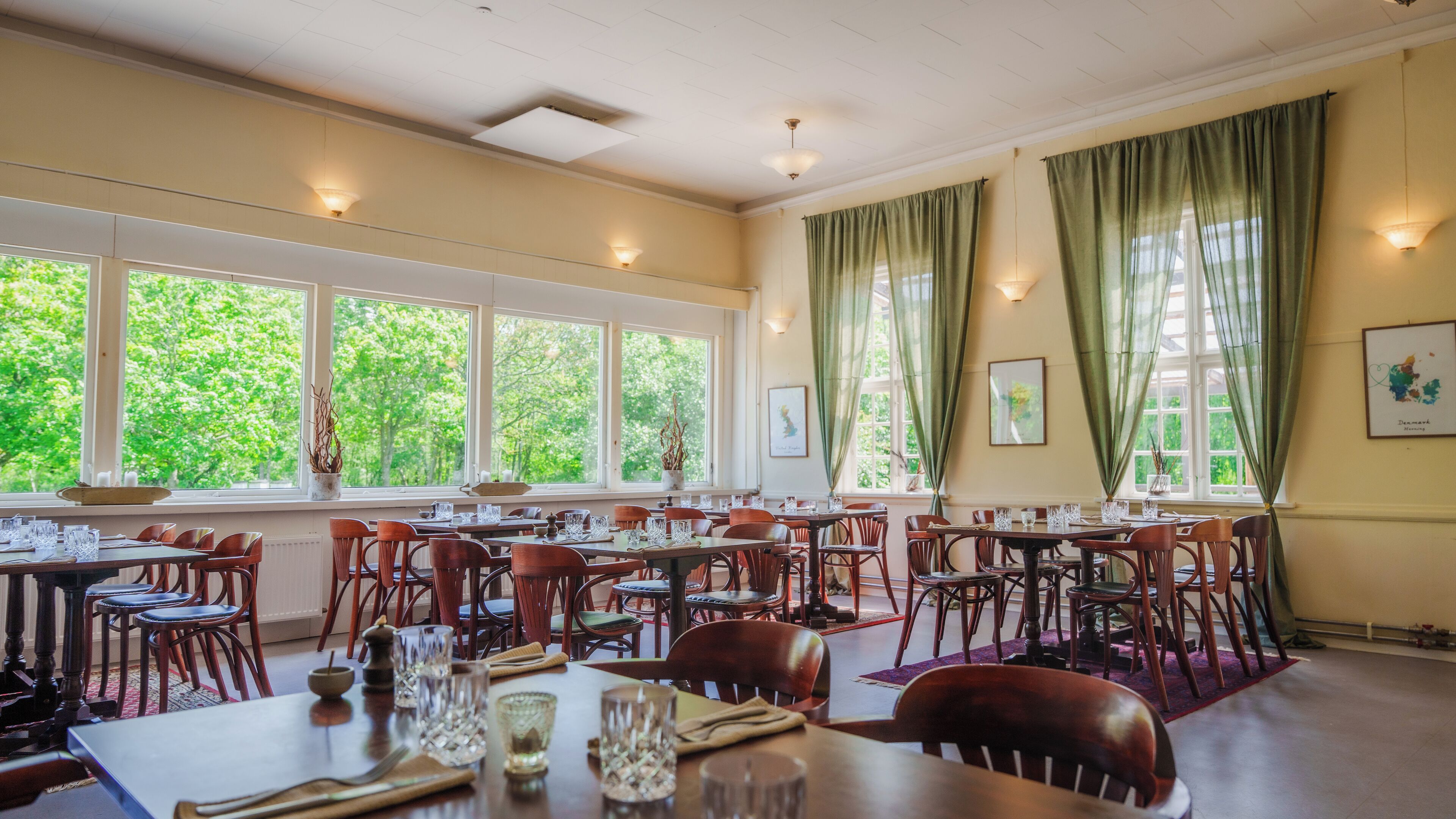 A dining area with multiple tables and chairs, large windows with curtains, and a view of greenery outside.
