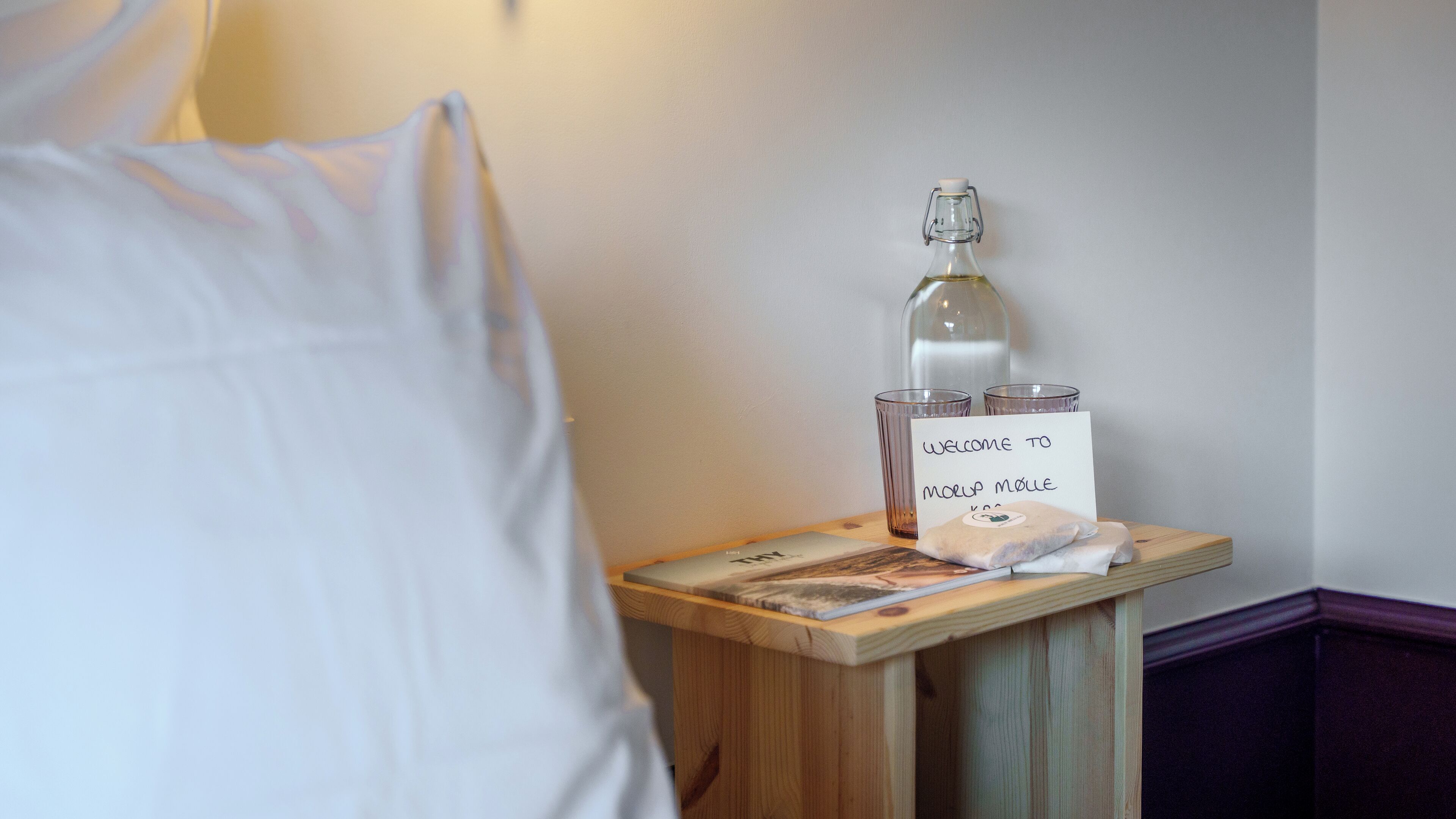 A neatly made bed with white linens, a bedside table with a glass bottle, two glasses, a welcome card, and a magazine.