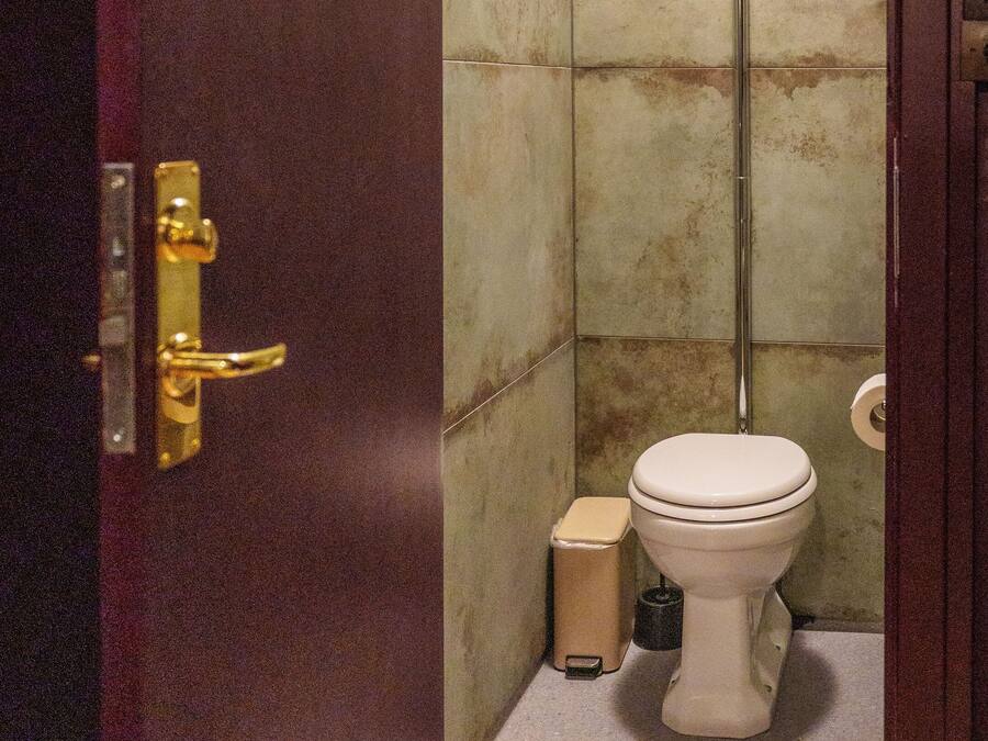 A small, narrow bathroom with a toilet, a shelf with toiletries, and a trash bin.