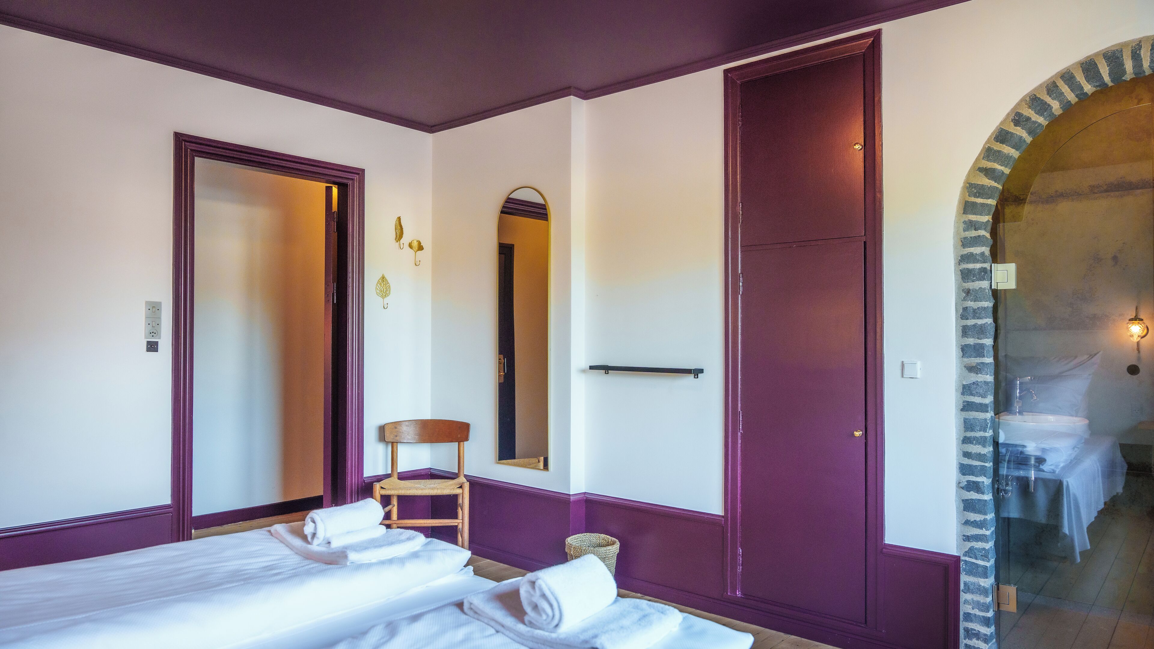 Habitación con dos camas, una silla, un espejo y un baño visible a través de un arco.