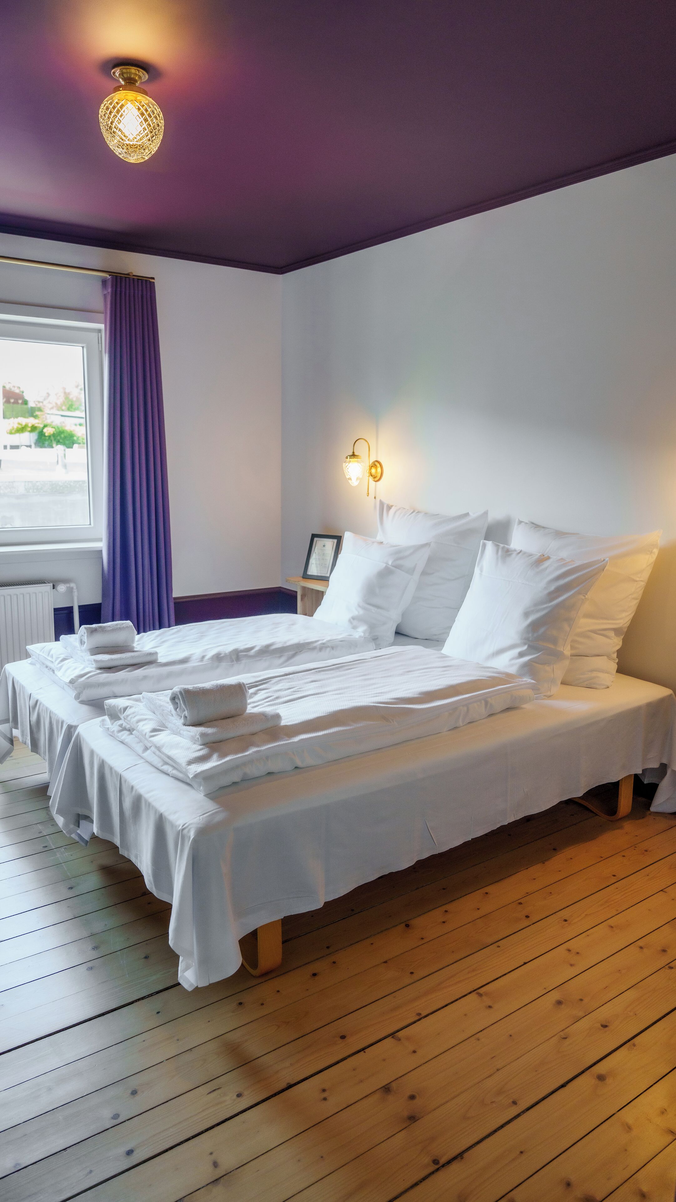 Un letto ben rifatto con lenzuola e cuscini bianchi, pavimento in legno, finestra con tende viola e un lampadario a parete.