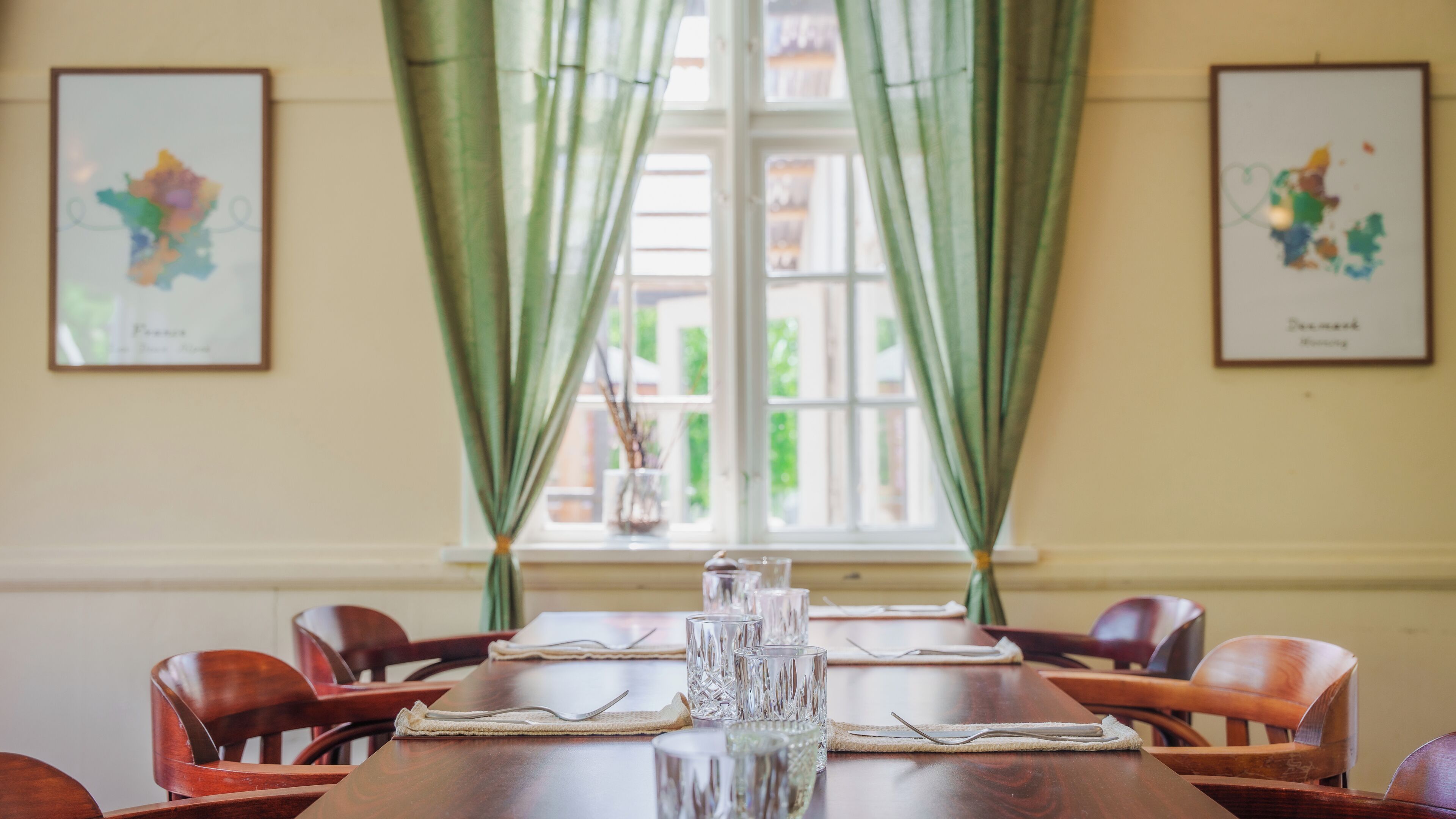 A dining table set with glassware and cutlery, flanked by chairs, with green curtains and framed artwork on the walls.