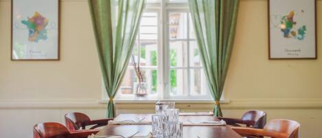 A dining table set with glassware and cutlery, flanked by chairs, with green curtains and framed artwork on the walls.