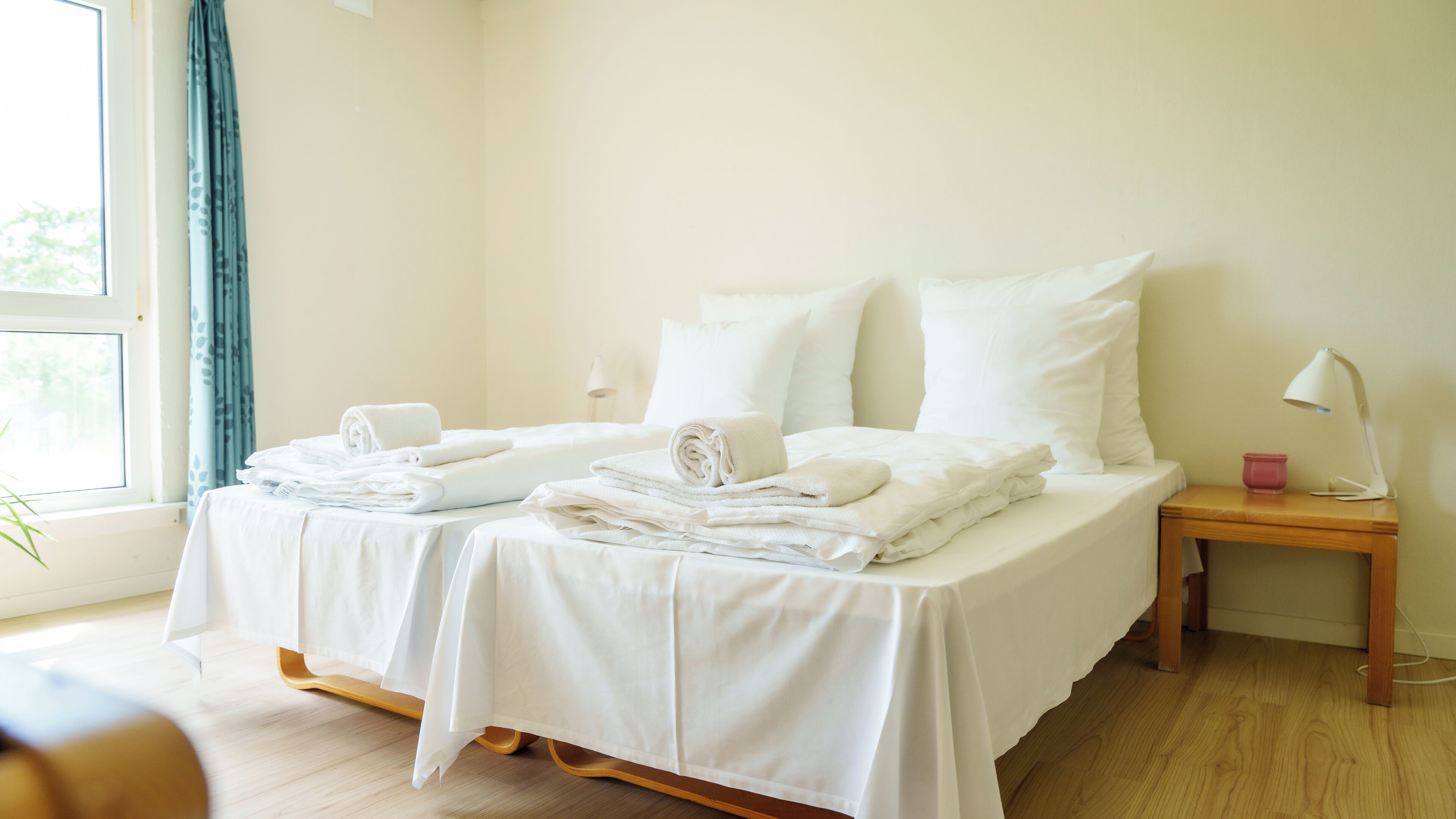 Una habitación de hotel limpia y minimalista con dos camas, ropa de cama blanca, una mesita de noche de madera y una ventana con cortinas.