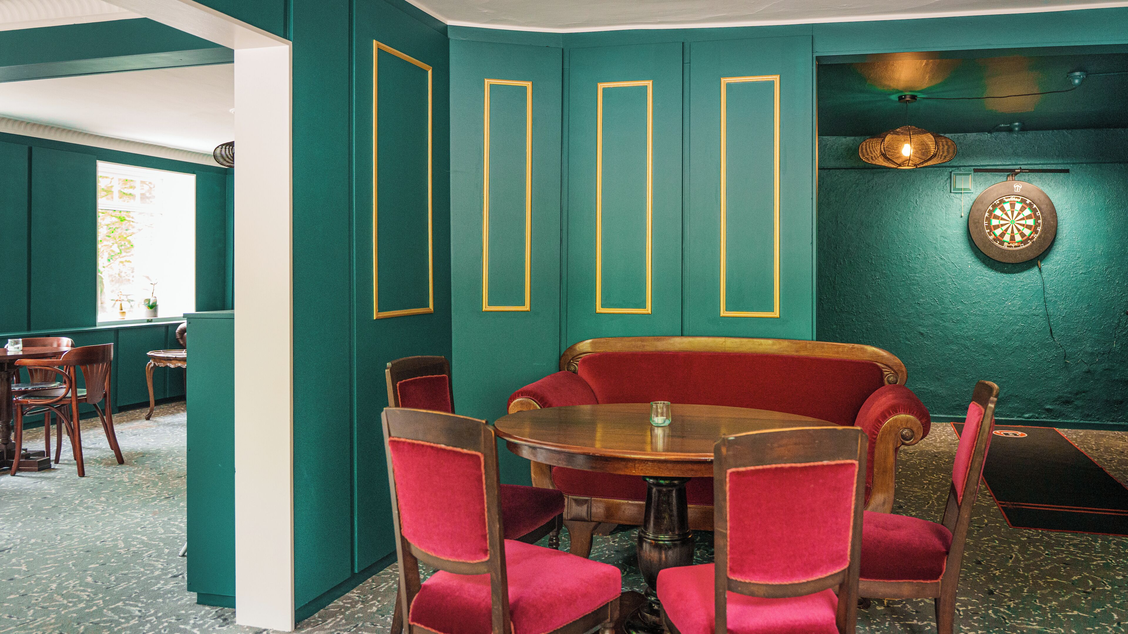 Un comedor con una mesa redonda y sillas tapizadas en rojo, paredes verdes y un dardo en la pared.