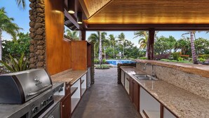An outdoor kitchen with a grill, sink, and wooden cabinets, overlooking a pool and tropical landscaping.