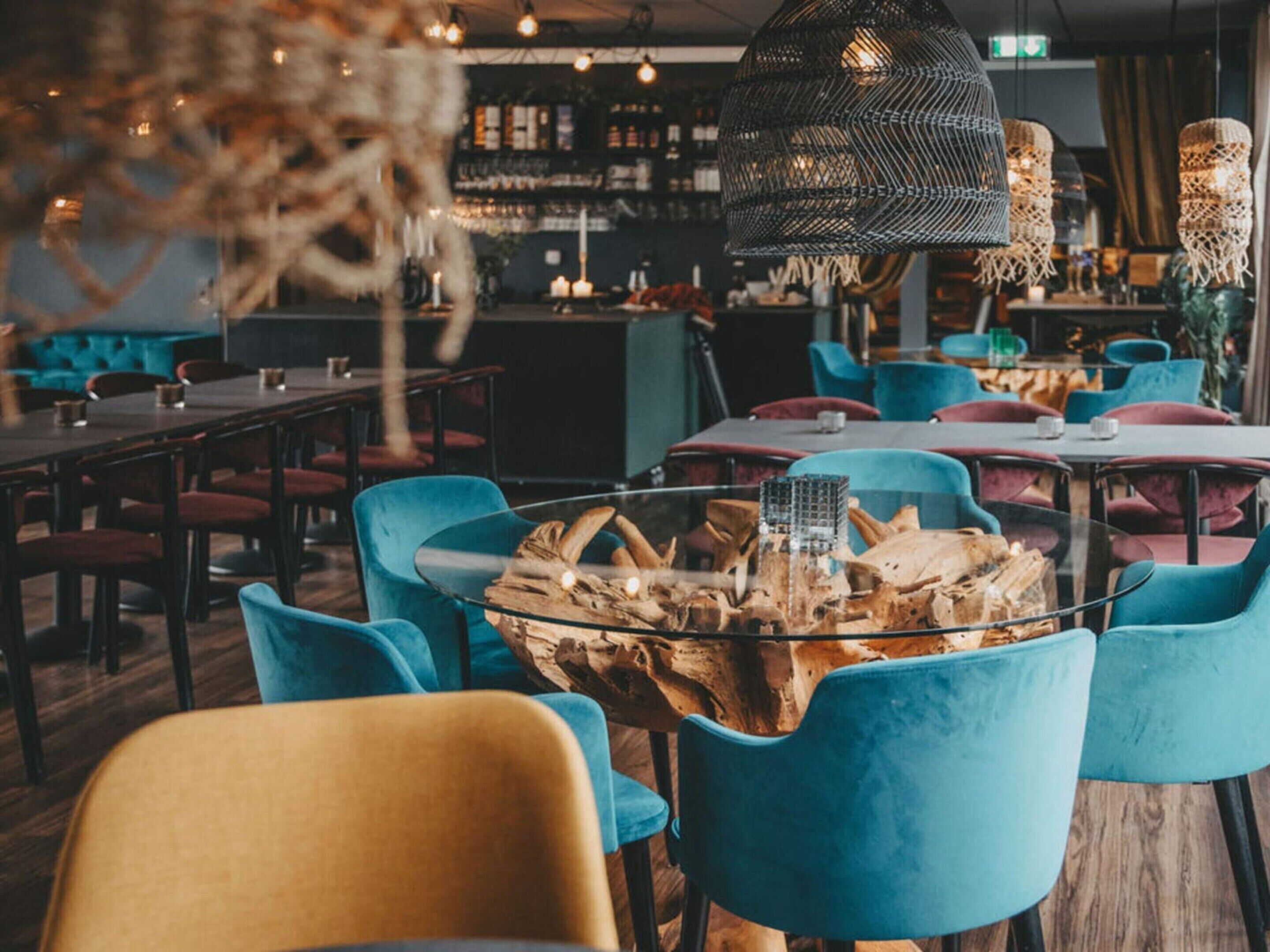Ein Restaurantinneres mit runden Tischen, blauen Stühlen und einem zentral platzierten Glastisch mit einer Holzdekoration.