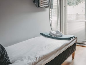Una cama individual con colchón blanco y cabecera oscura, una toalla doblada sobre la cama, y una ventana con persianas entreabiertas.