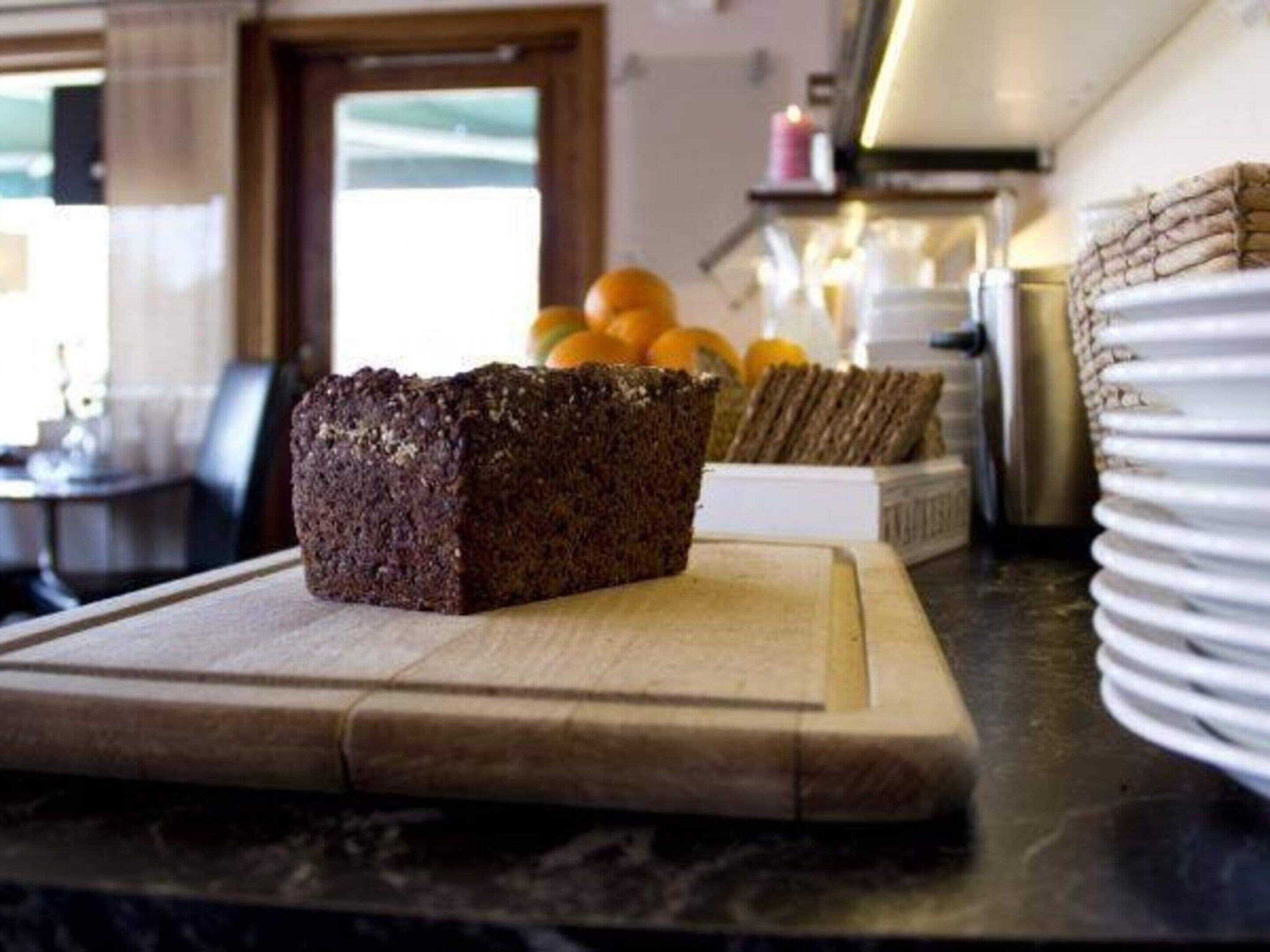 A kitchen counter with a loaf of dark bread on a wooden cutting board, a stack of white plates, and a basket of oranges in the background.