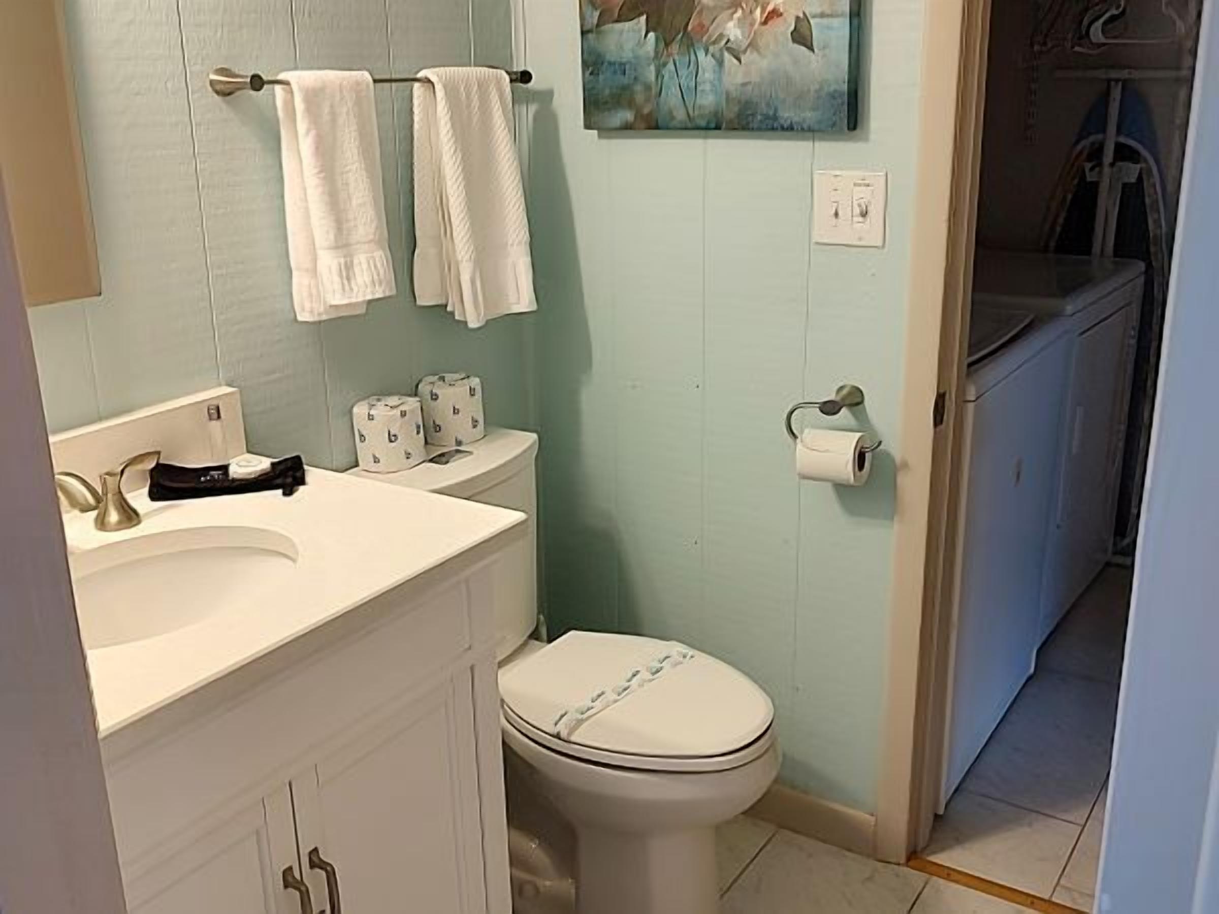 A bathroom with a white sink, two towels hanging on a towel bar, a toilet, and a painting on the wall.