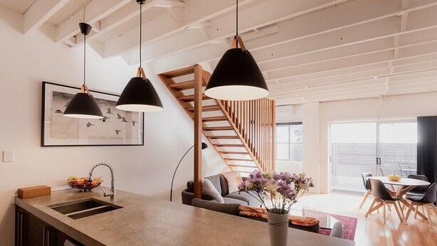 A modern kitchen with a central island, pendant lights, a wooden staircase, and a dining area with a view. - Urban Warehouse Retreat in West Hobart (West Hobart)
