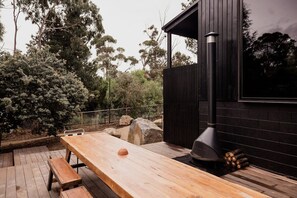 An outdoor wooden deck with a table and benches, a black building with a chimney, and surrounding trees. - 'Sisters' - Cliff top cabin with hot tub (Spring Beach)