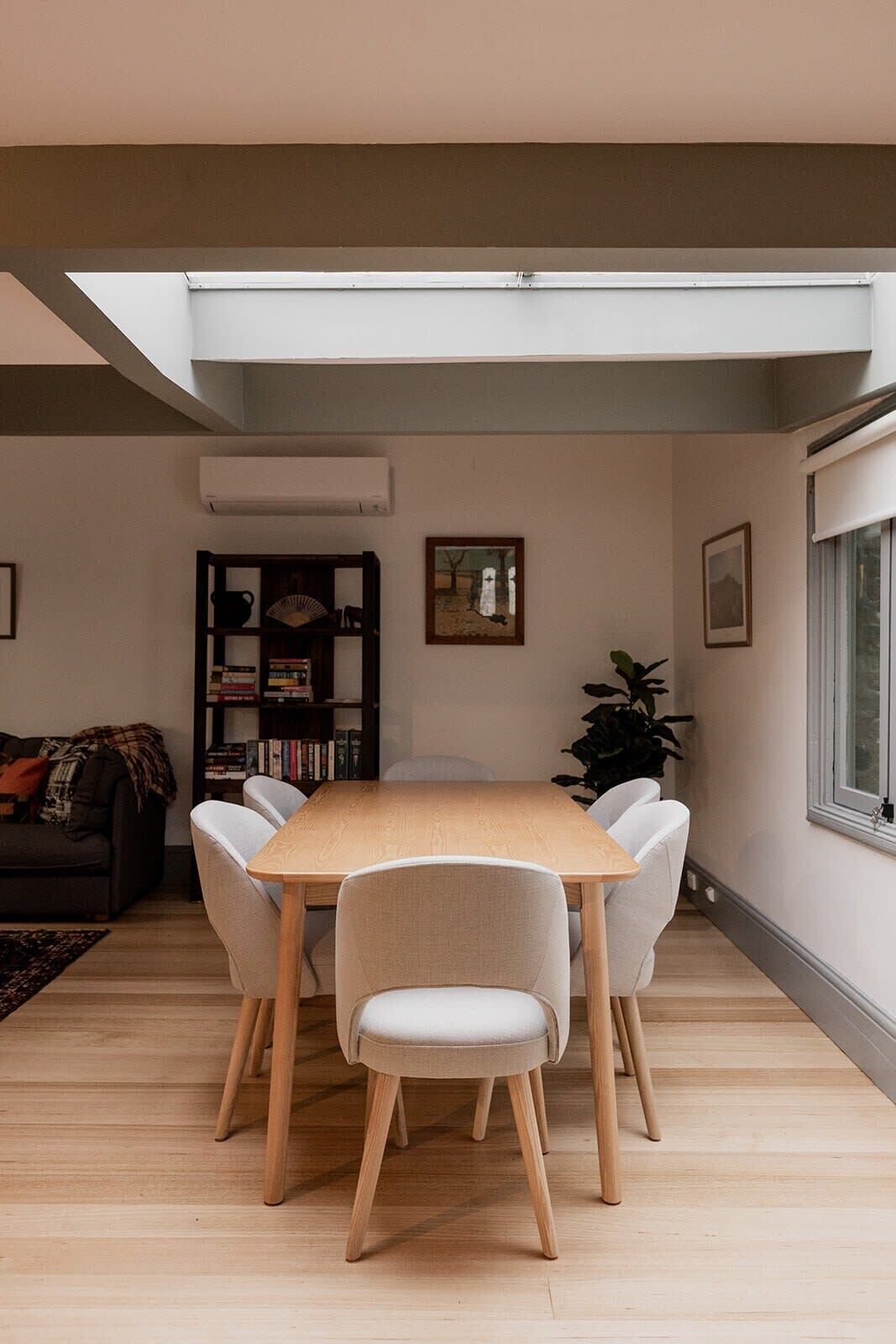 A dining area with a wooden table and four chairs, a bookshelf, a plant, and a window with blinds.