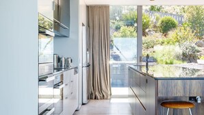 A modern kitchen with a large window, a dining area with a bar stool, and a view of a garden. - Architectural Glass House with Breathtaking Ocean Views (Taroona)