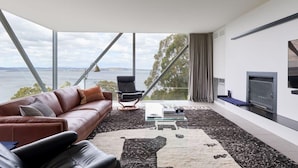 A modern living room with a large window, a leather sofa, a coffee table, and a fireplace. - Architectural Glass House with Breathtaking Ocean Views (Taroona)