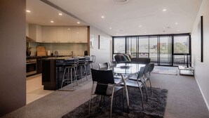 A modern kitchen with a dining area, featuring a glass table and bar stools, a large window with a city view, and recessed ceiling lighting. - Fantastic CBD Apartment with Views (Hobart)