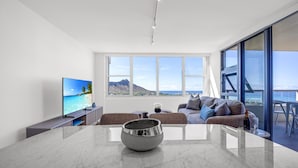 A modern living room with a large window, a flat-screen TV, a marble coffee table, and a sofa with cushions.