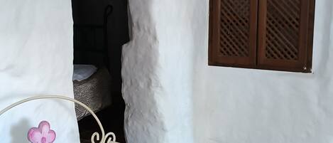 A room with a white wall, a wooden door, a window with wooden shutters, a white bench with a pink flower design, and terracotta floor tiles.