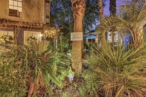 A well-maintained garden with a palm tree, a sign reading 'Welcome to the Garden,' and a building with a rustic facade.