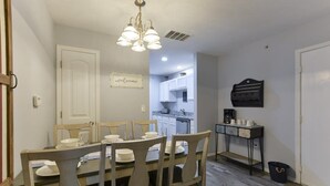 A dining area with a wooden table and chairs, a chandelier, and a kitchen in the background.