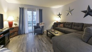 A living room with a sofa, armchair, and coffee table. There is a television, a lamp, and a window with curtains.