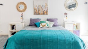 A neatly arranged bedroom with a teal bedspread, a round mirror, and a wall art piece.
