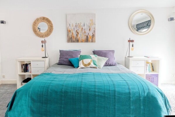 Un dormitorio ordenado con una colcha color aguamarina, un espejo redondo y una obra de arte en la pared.