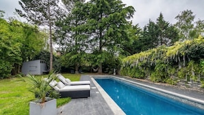 A rectangular swimming pool with a tiled deck, surrounded by lush greenery and trees.