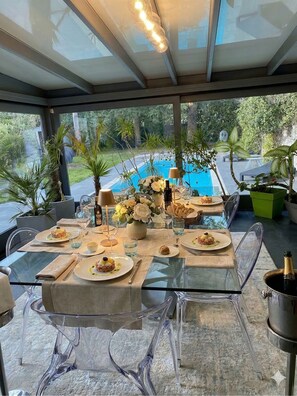 An outdoor dining area with a glass table, chairs, and a view of a swimming pool.