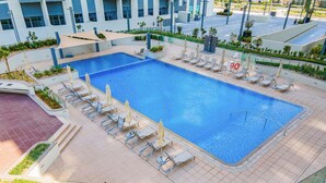 A large rectangular pool with multiple lounge chairs and umbrellas, surrounded by a tiled deck and greenery.