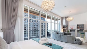Et moderne soveværelse med en stor seng, en grå sofa og udsigt over byens skyline gennem gulv til loft-vinduer.