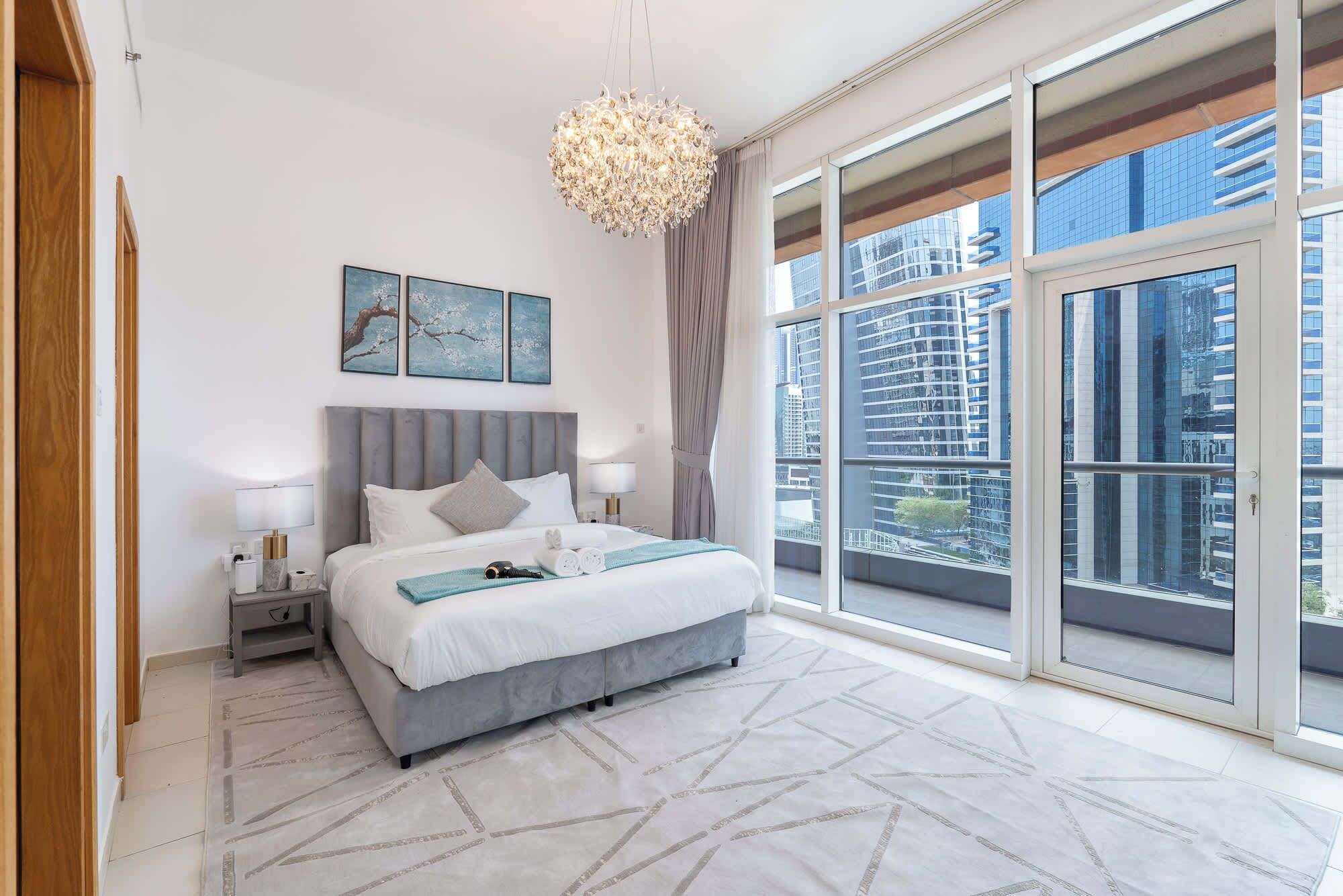 Une chambre d’hôtel moderne avec un grand lit, un lustre, des œuvres d’art accrochées au mur et une vue sur la ville grâce à de grandes fenêtres.