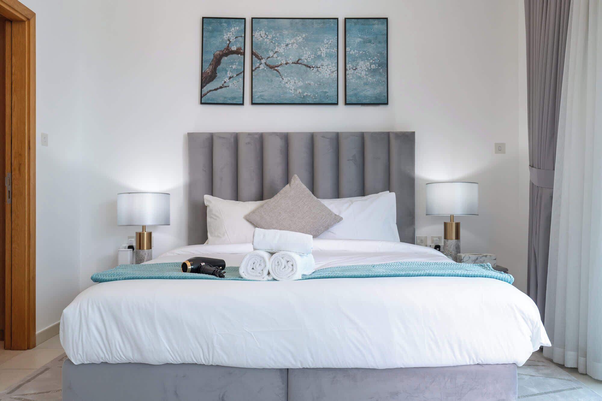 A neatly made bed with a grey headboard, two bedside lamps, and a pair of rolled towels on the bed.