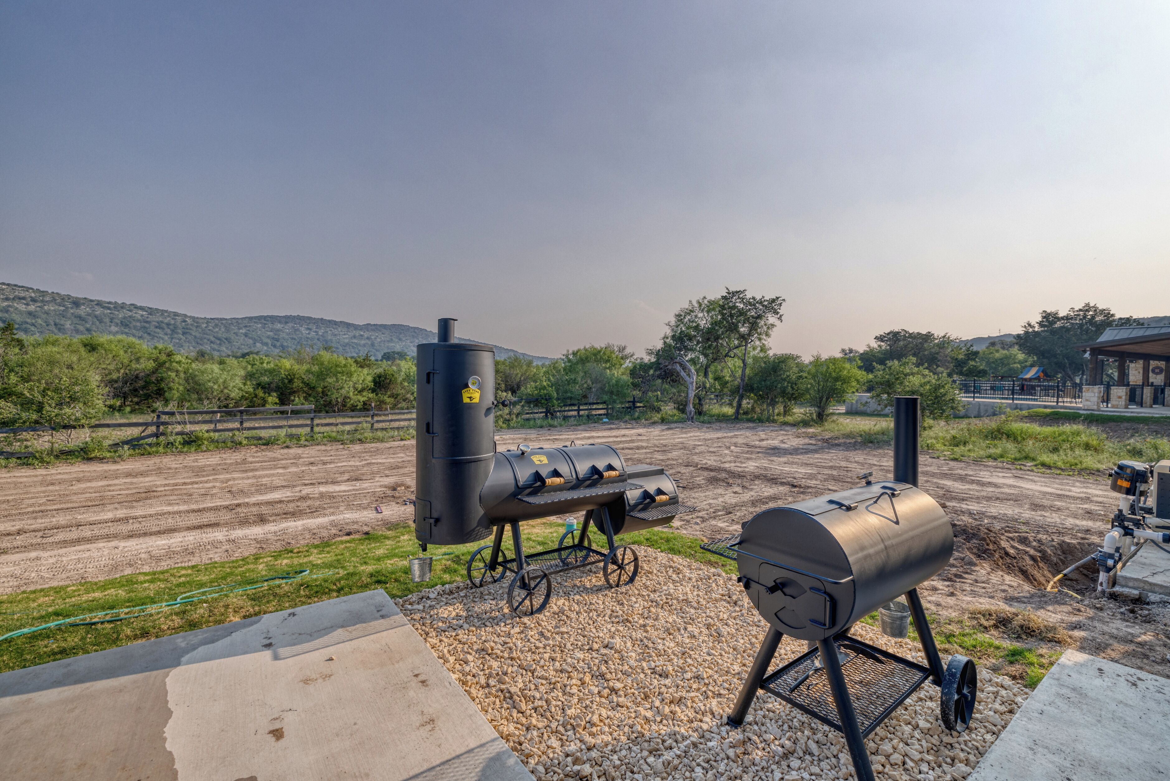 Un espacio para barbacoa con una parrilla y un ahumador, rodeado de grava y un sendero de tierra, con una cerca y árboles al fondo.