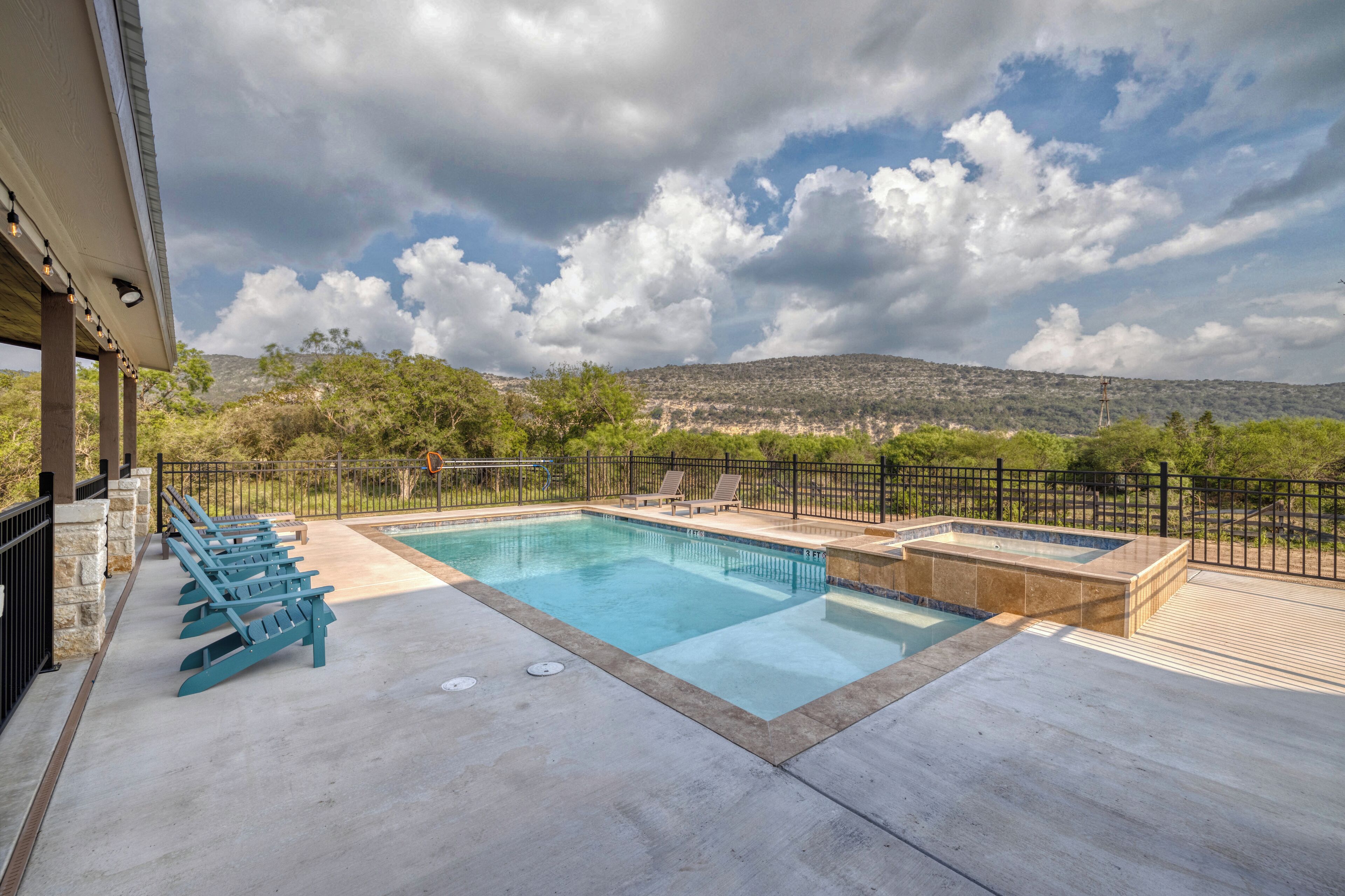 Une piscine dotée de chaises longues et d’un jacuzzi, entourée d’une clôture et d’arbres, avec une chaîne de montagnes en toile de fond.