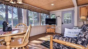 A cozy cabin interior with a dining area, a seating area with a TV, and large windows offering a view of trees and a lake. - Stepaway Vacation Rentals-Cozy Cottage (St.Germain)