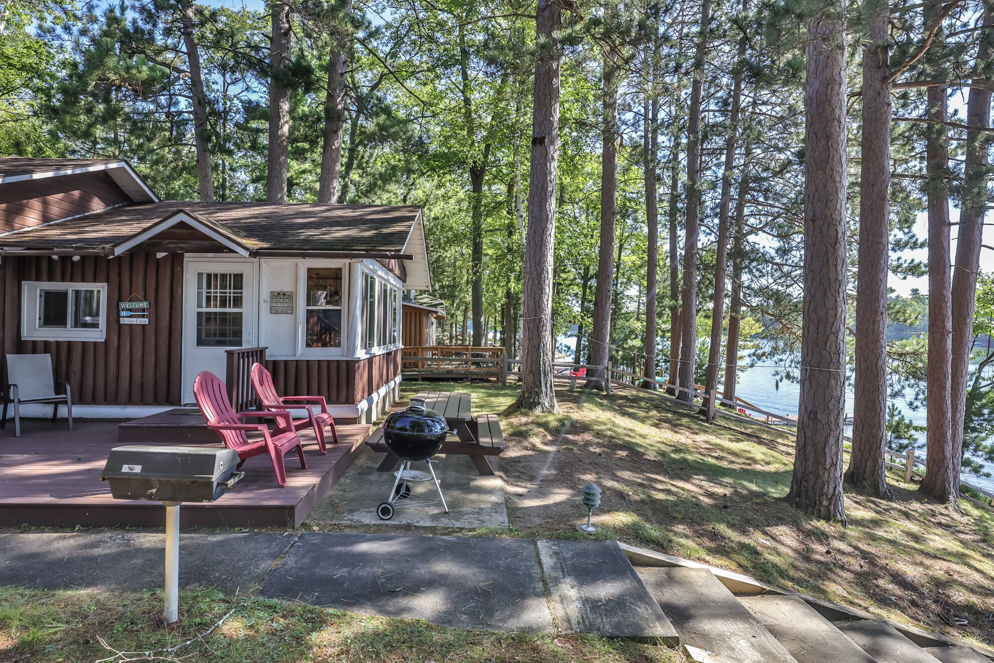 Una cabana amb un porxo, cadires Adirondack vermelles, una barbacoa i vistes a un llac envoltat d'alzines altes.