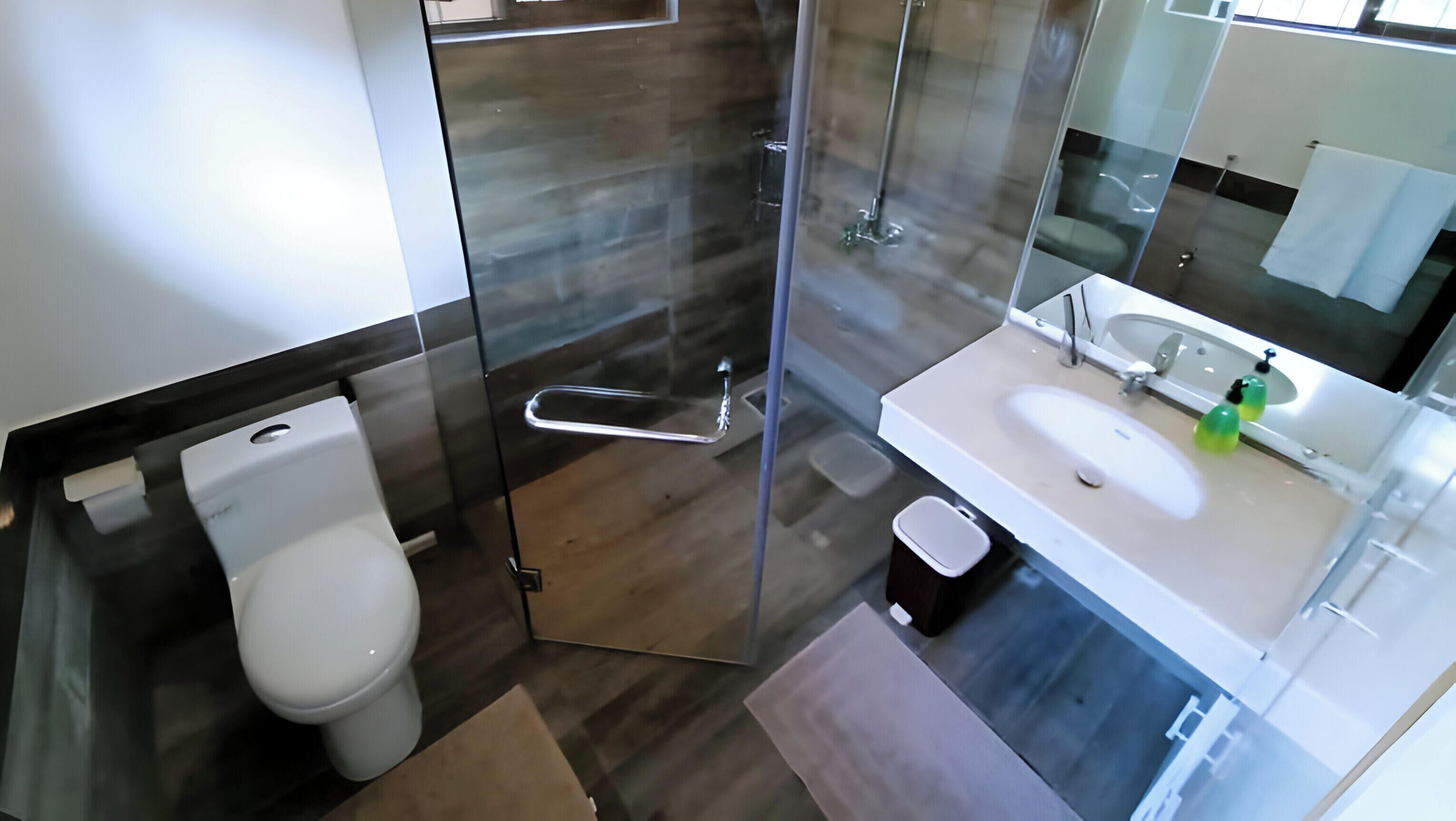 Un baño moderno con cabina de ducha de vidrio, inodoro, lavabo con espejo y un cubo de basura pequeño.