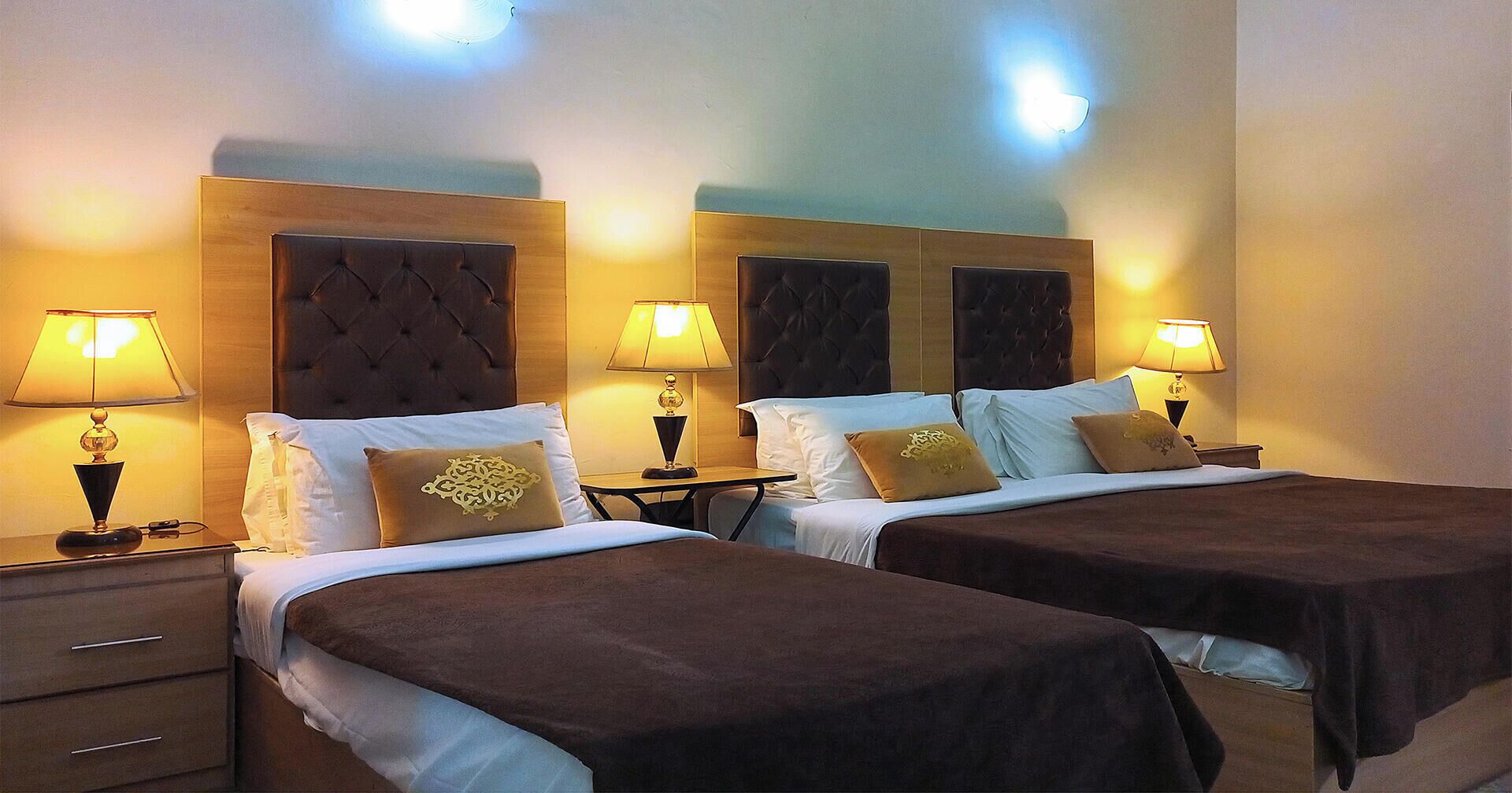 A hotel room with two beds, each with a headboard, bedside tables with lamps, and a small table between the beds.