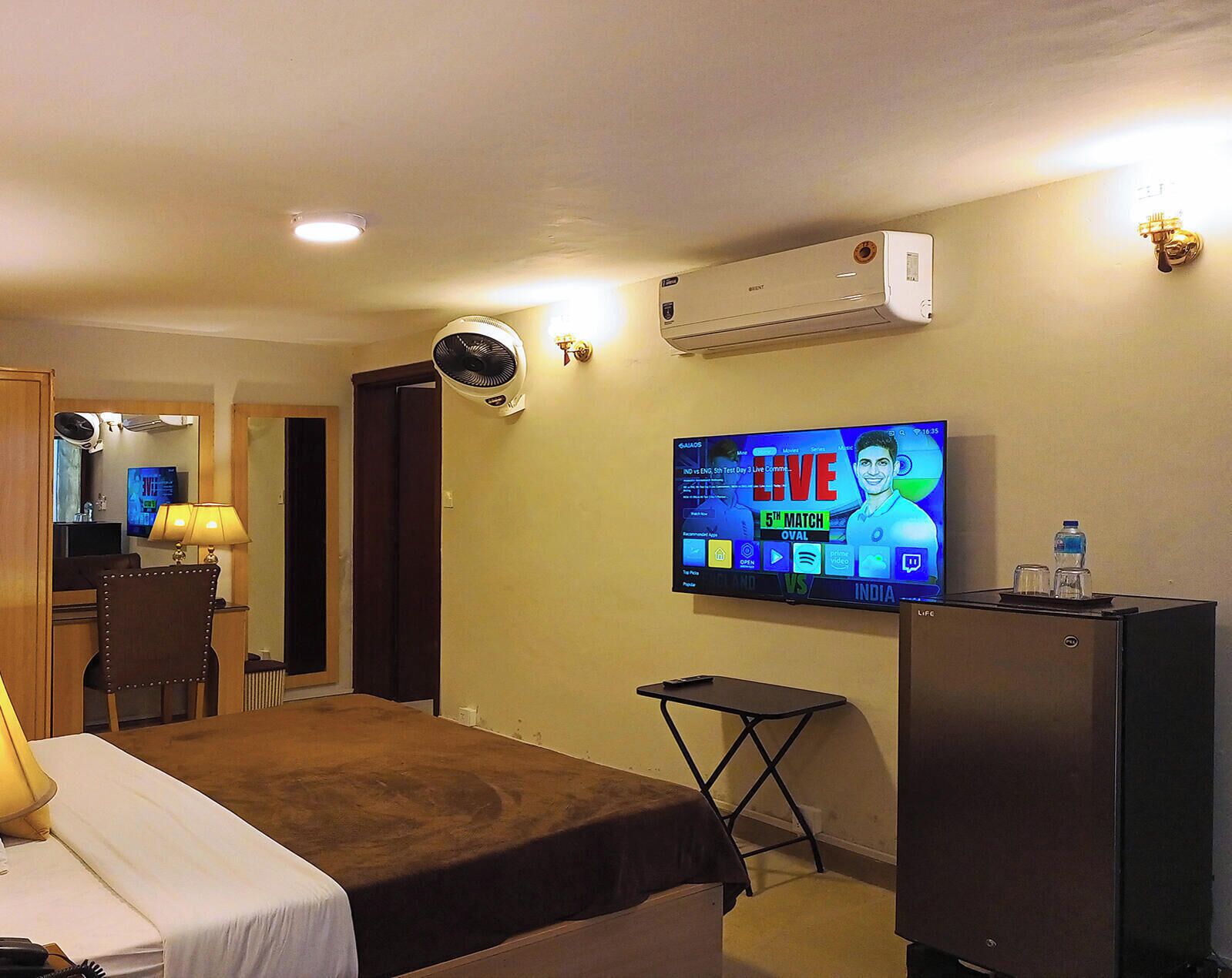 A hotel room with a bed, a desk, a chair, a TV mounted on the wall, an air conditioner, and a water bottle on the desk.