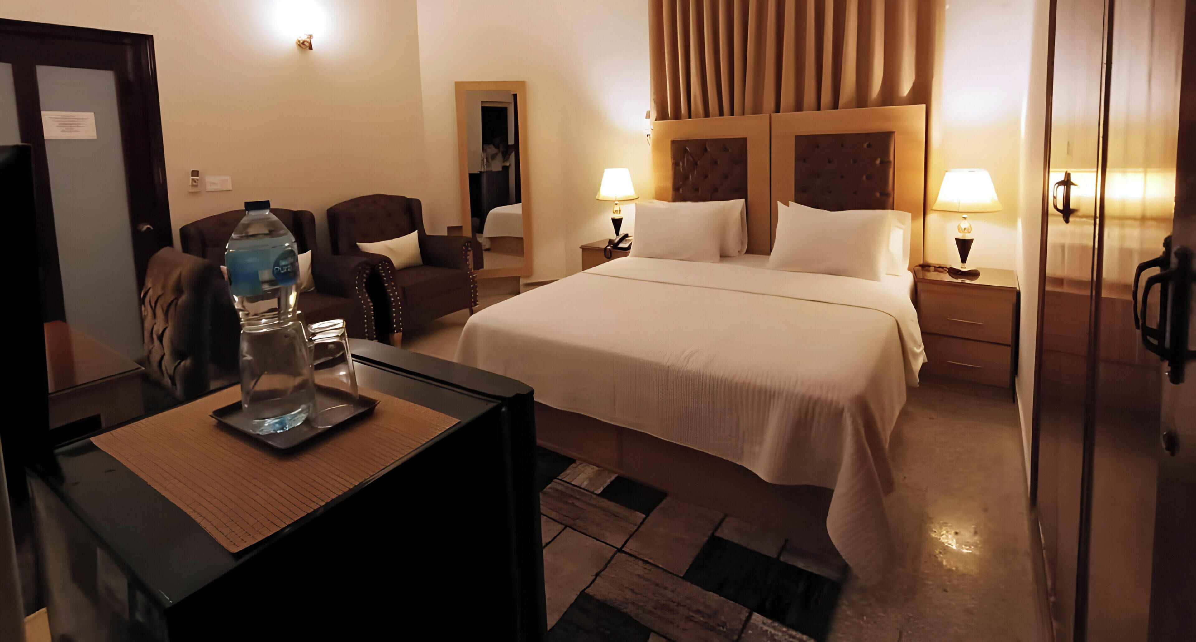 Quarto de hotel com uma cama, duas mesas de cabeceira, uma escrivaninha com cadeira, um espelho, um ventilador de teto e um ar condicionado.