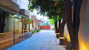Un sendero bordeado de árboles y plantas en macetas, que conduce a un edificio con fachada de vidrio y un logotipo visible.