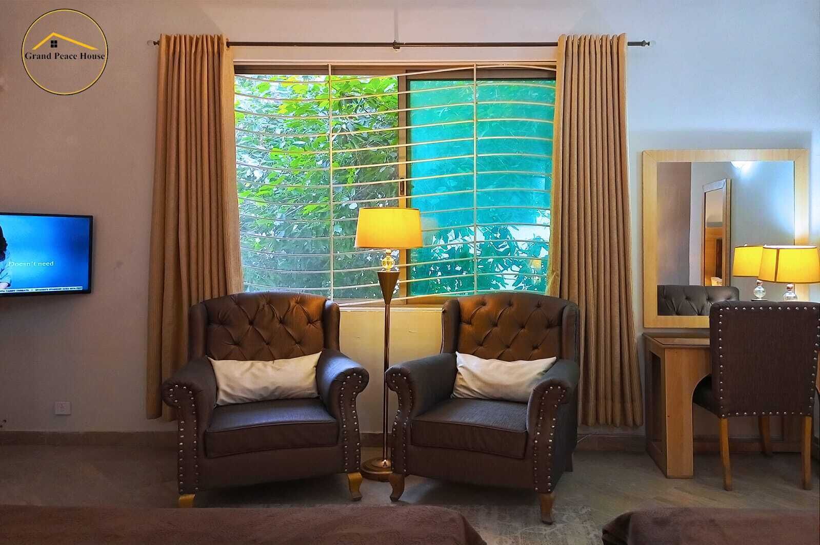 A hotel room with two leather armchairs, a floor lamp, a television, a large window with curtains, and a wooden desk with a mirror.