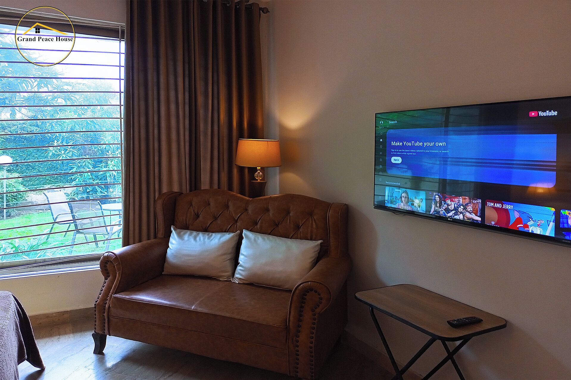 A room with a brown leather sofa, a small table, a TV on the wall, and a window with curtains.