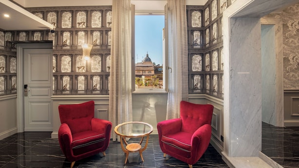 A luxurious room with two red armchairs, a glass table, and a view of a historic building through a large window. - The Inn at the Spanish Steps Elegant Collection - The Jewel (Rome)