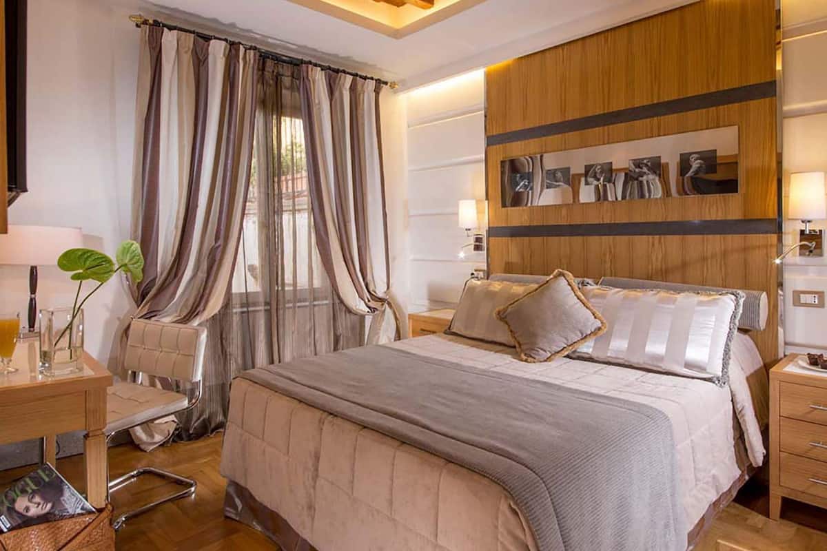 A modern hotel room with a large bed, a wooden headboard, a desk with a lamp, and a bedside table with a glass of orange juice.