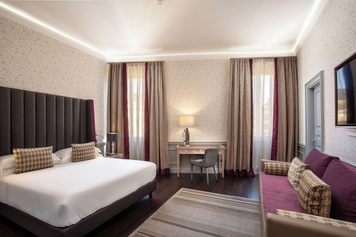A hotel room with a large bed, a desk, a chair, a sofa, and a TV.